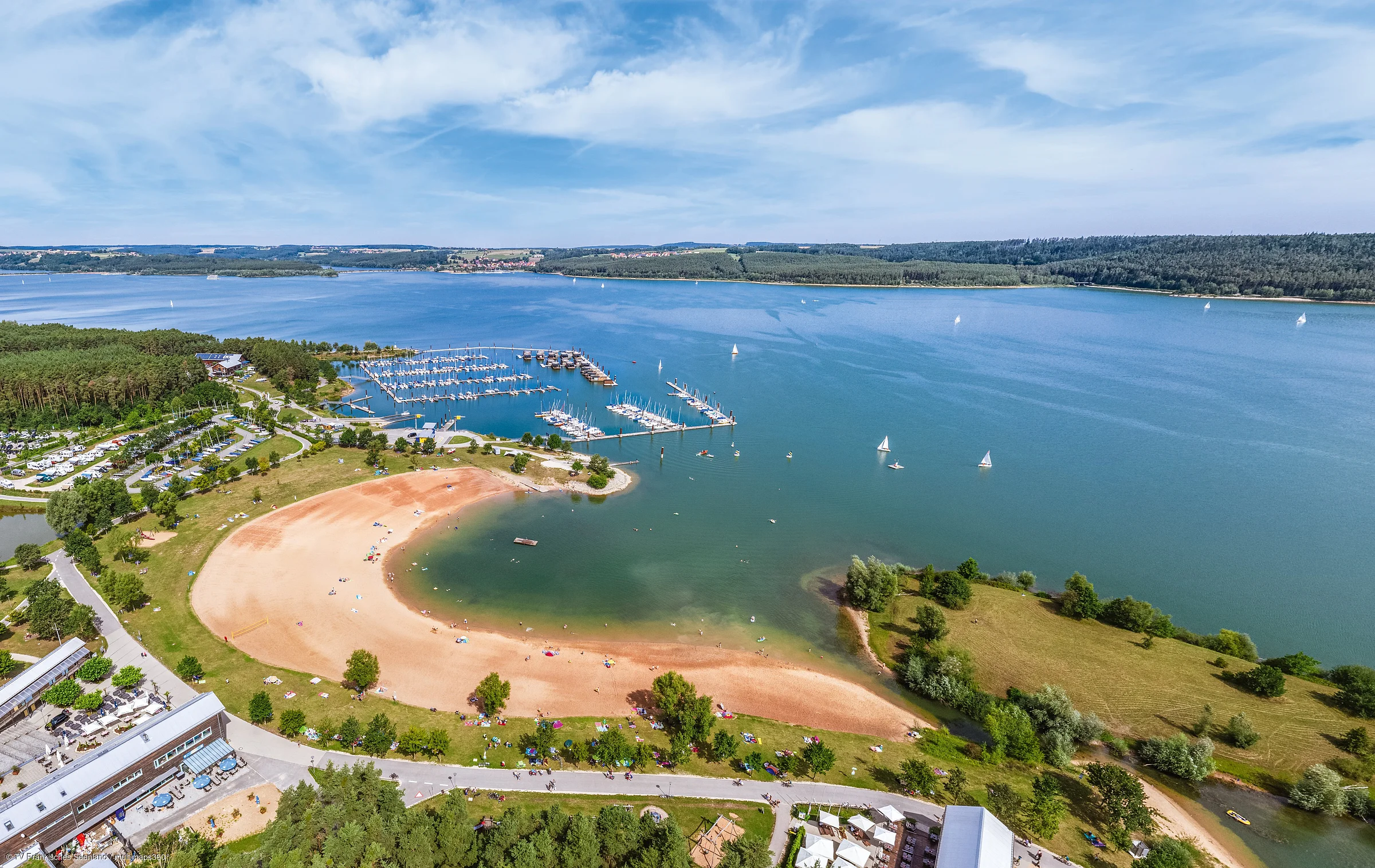 Luftaufnahme eines Sandstrands mit Liegewiese, Bootshafen und Segelbooten auf einem großen See unter blauem Himmel.