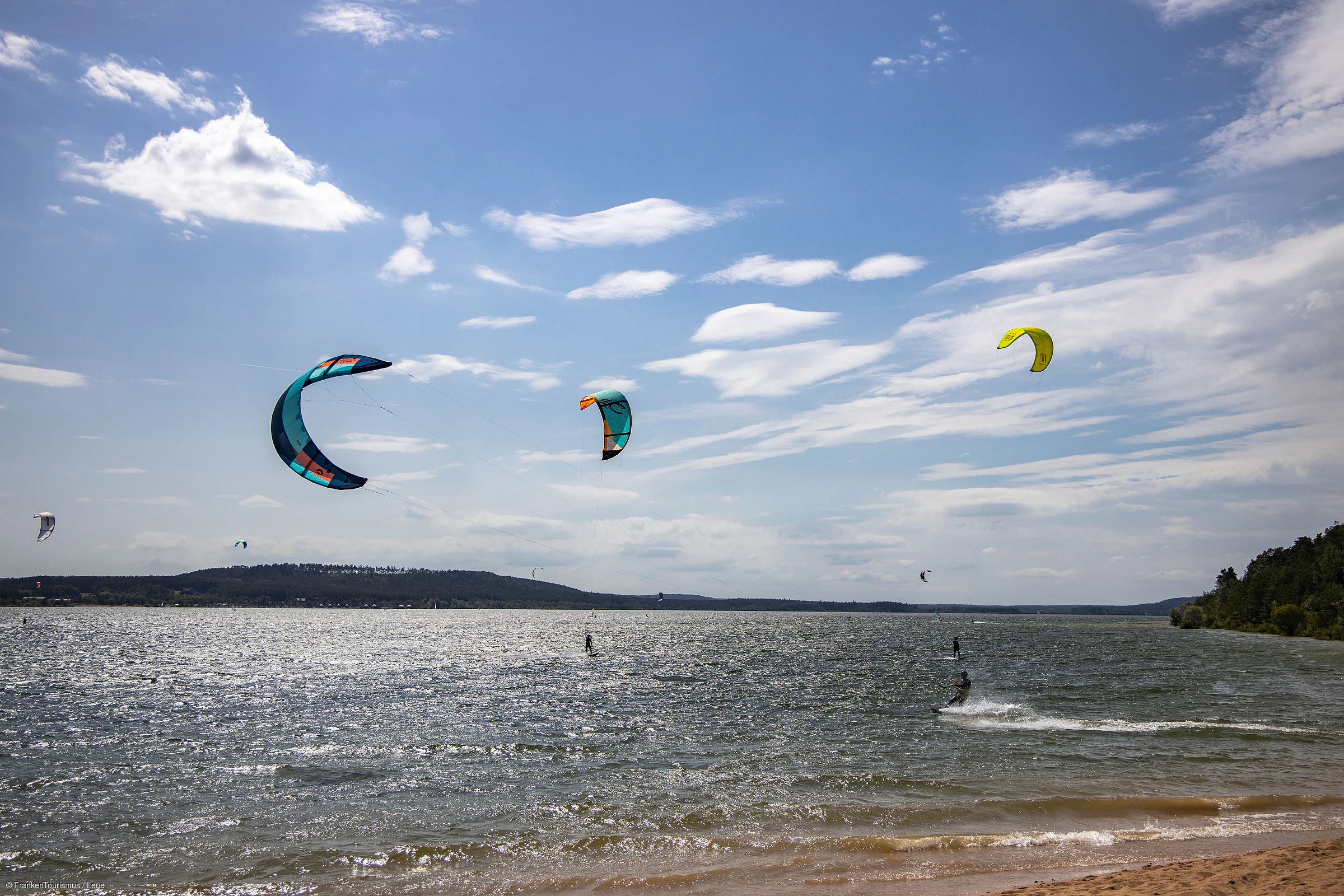 Kitesurfer mit bunten Lenkdrachen auf einem See bei leicht bewölktem Himmel und Sandstrand im Vordergrund.