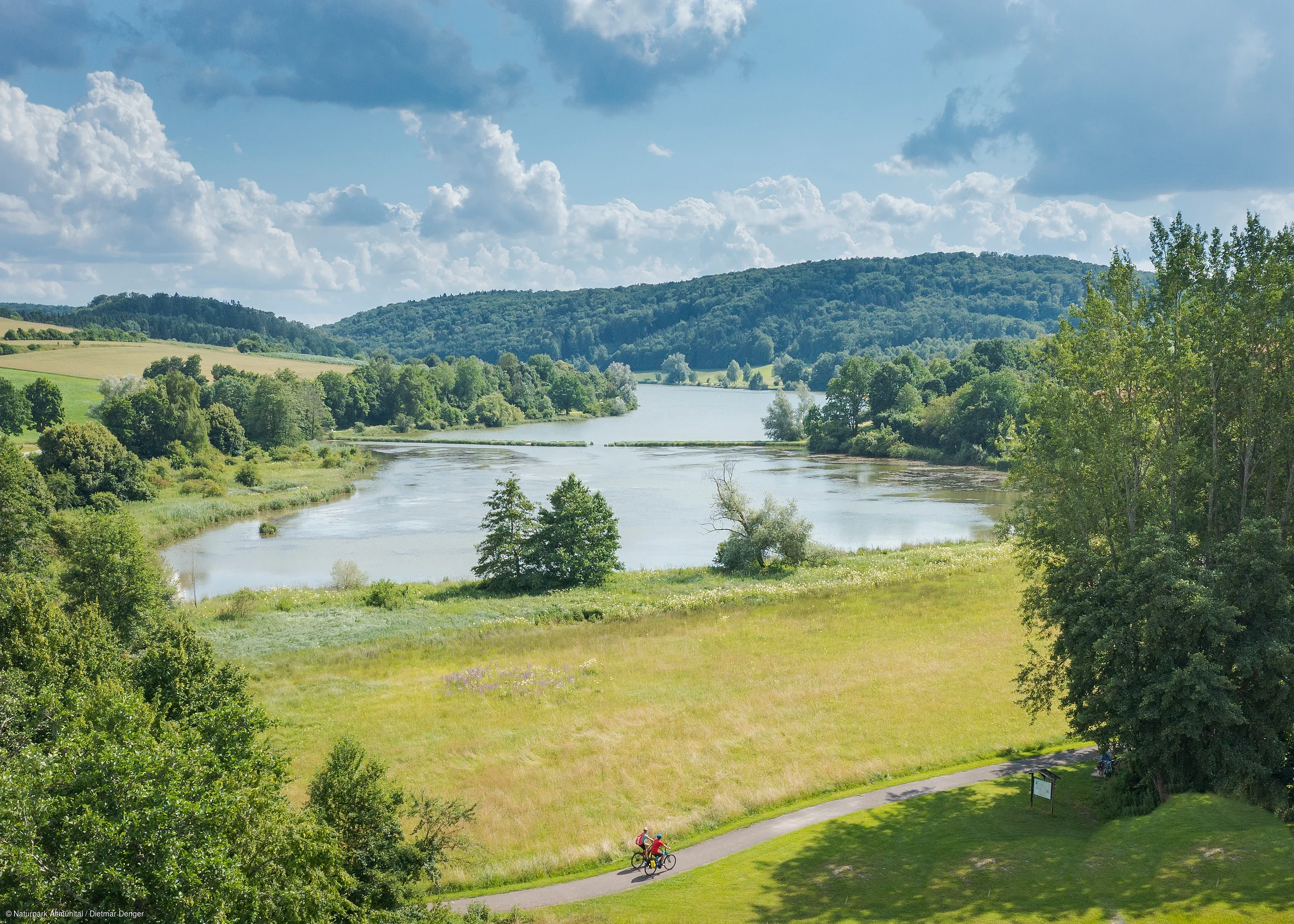 Landschaft mit See, Wiesen, Wald und zwei Radfahrern auf einem Weg im Vordergrund bei bewölktem Himmel.