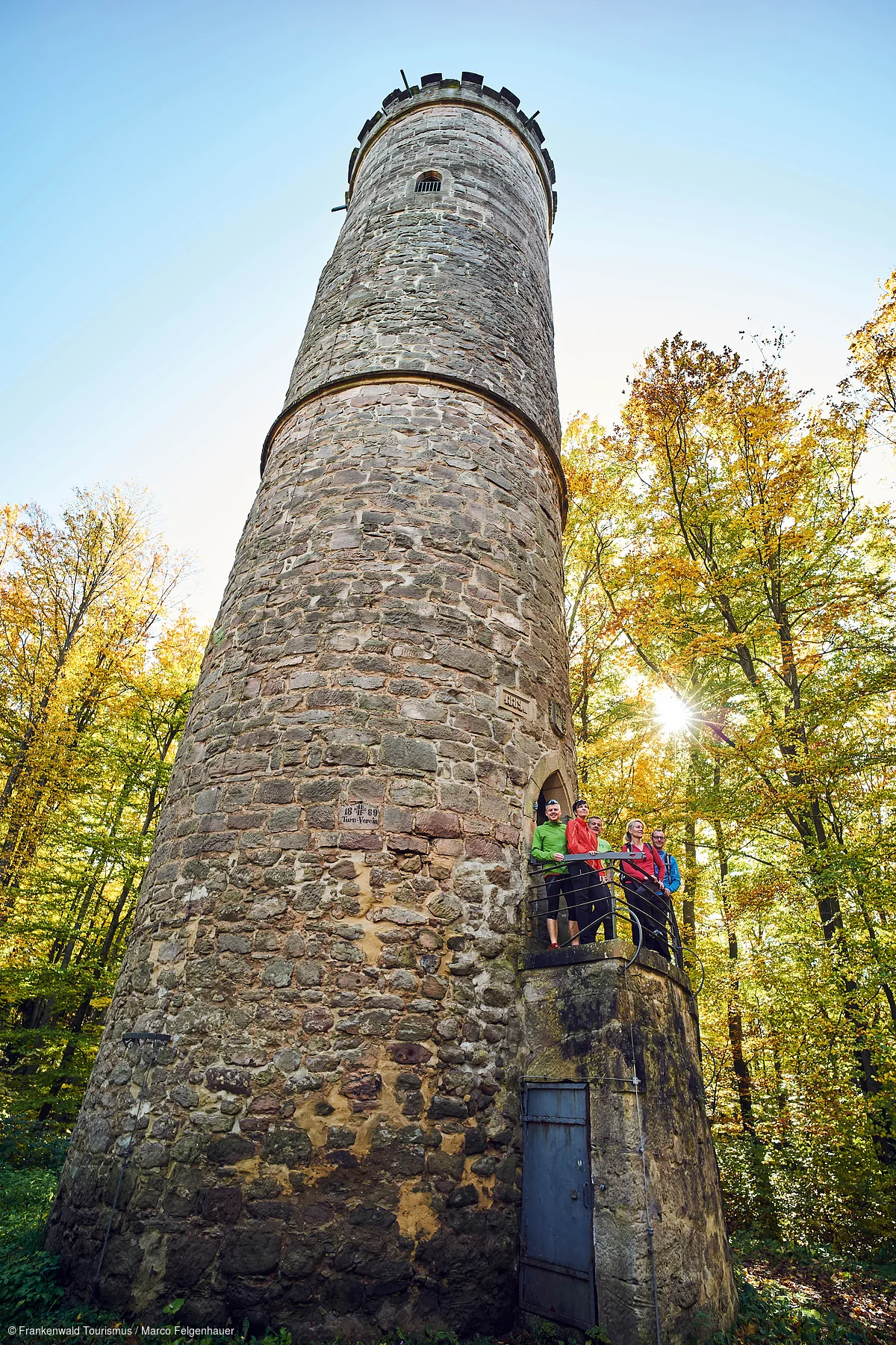 Steinturm mit Aussichtsplattform, auf der fünf Personen stehen, umgeben von herbstlichen Bäumen bei Sonnenschein.