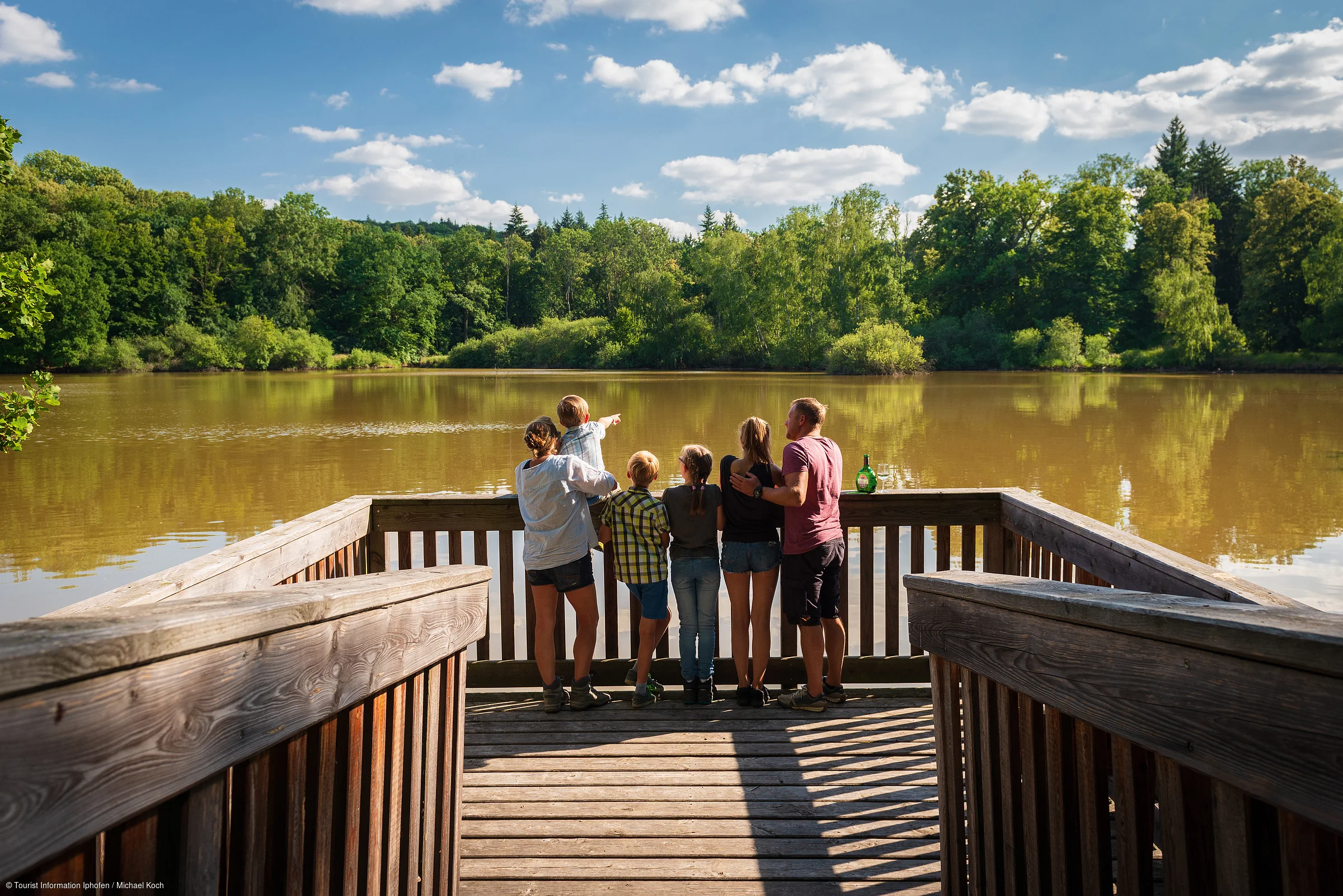 Sechs Personen stehen auf einem Holzsteg und blicken auf einen See mit bewaldetem Ufer bei sonnigem Himmel.