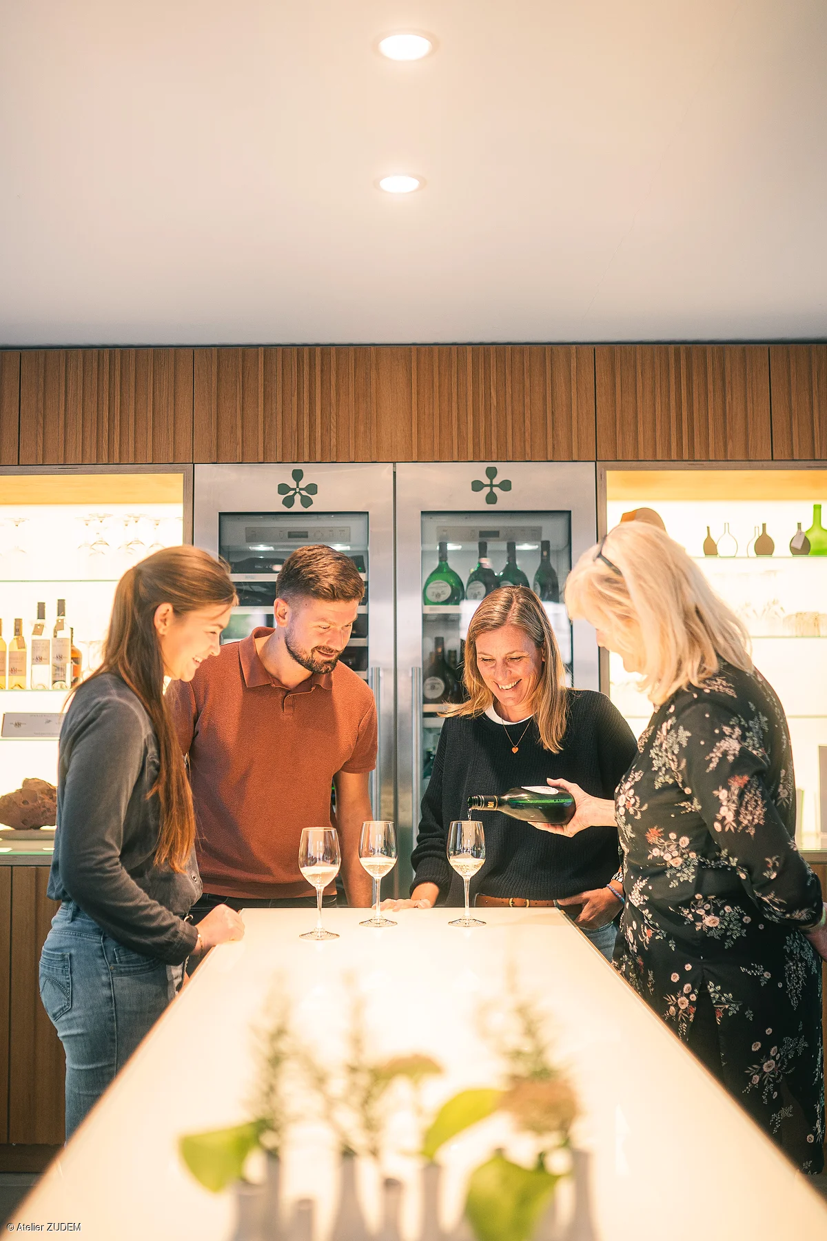 Vier Personen stehen an einem beleuchteten Tresen, eine gießt Wein in Gläser.