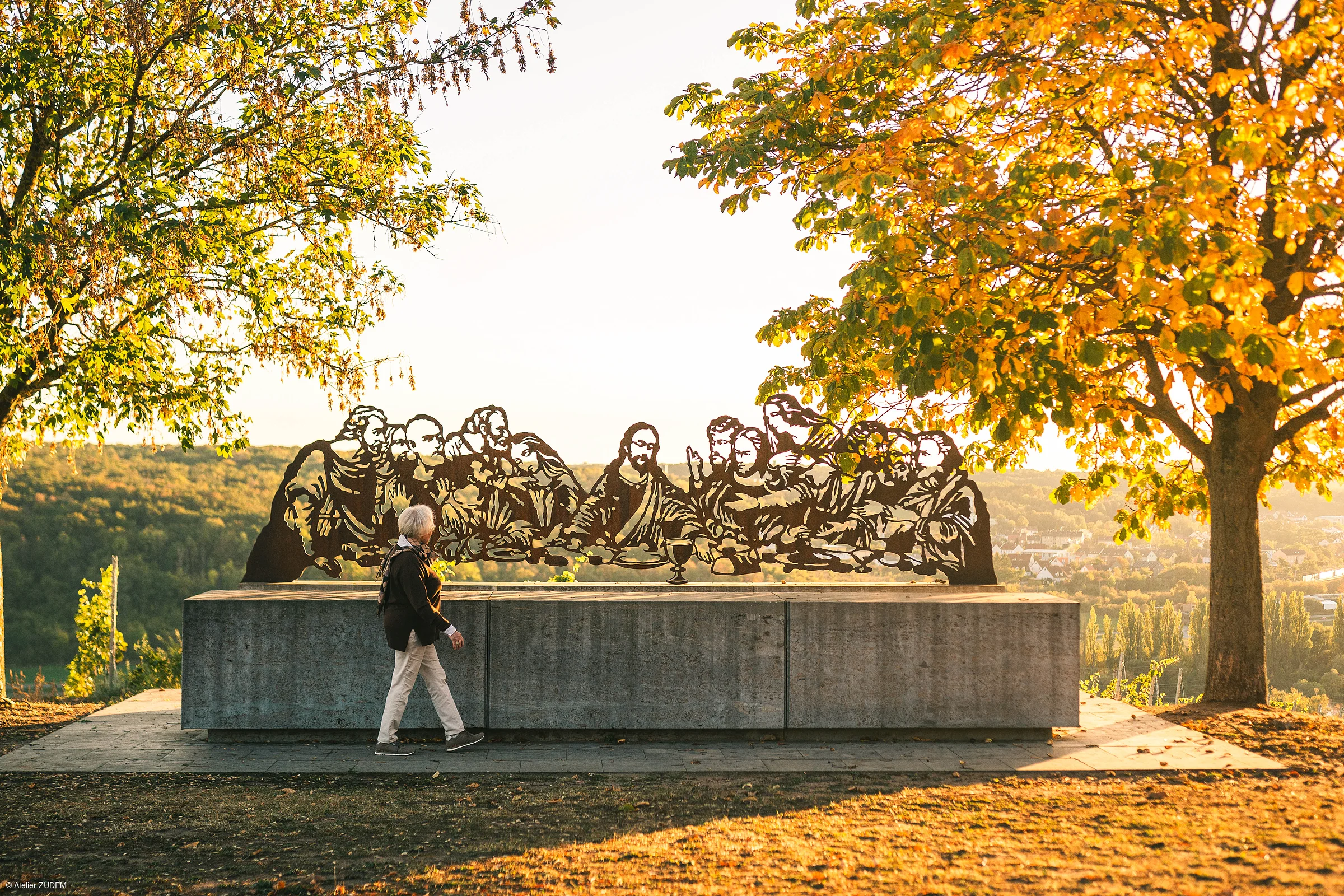Skulptur der letzten Abendmahl-Szene auf Betonpodest im Park mit herbstlichen Bäumen und Person im Vordergrund.