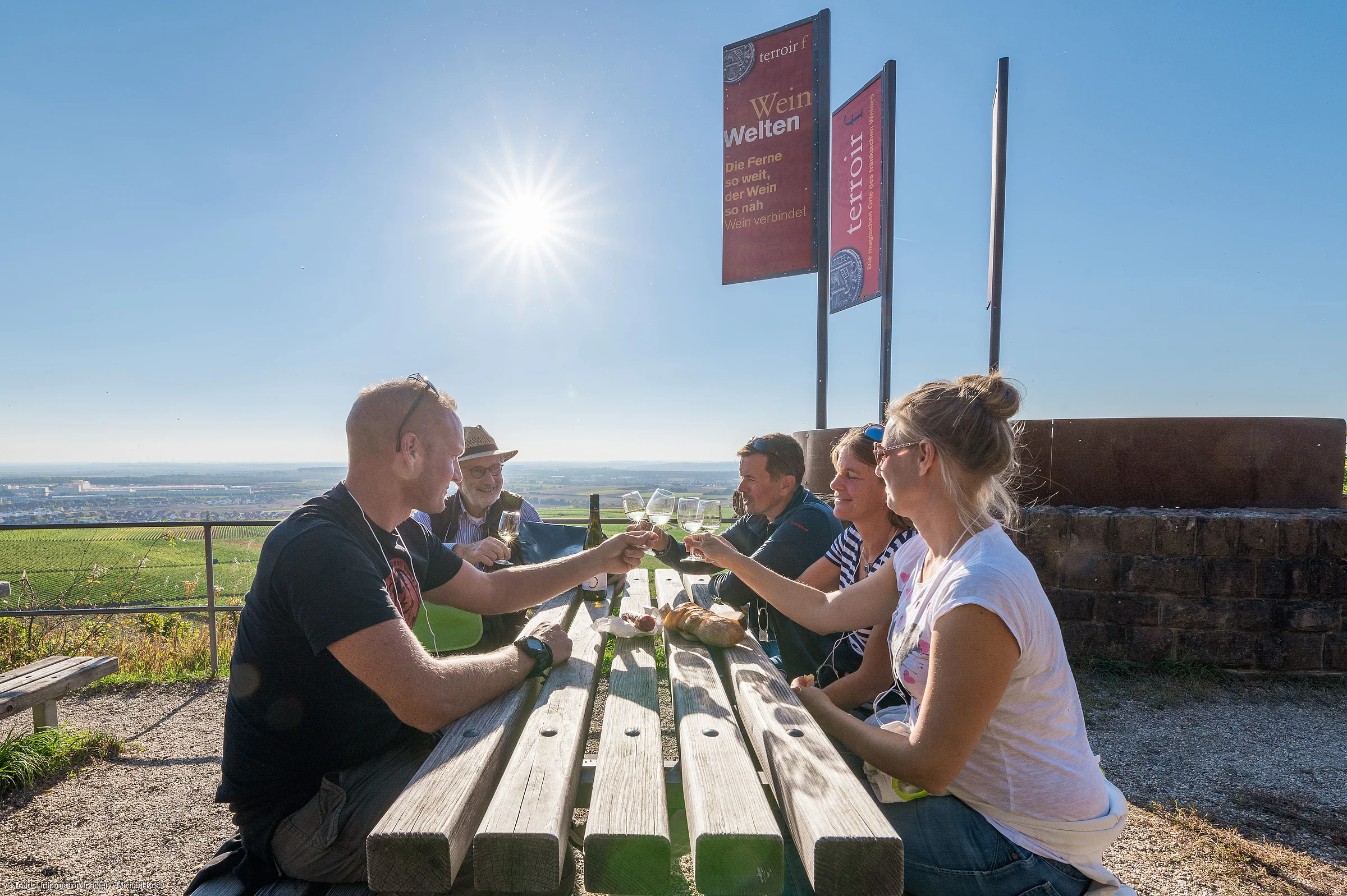 Fünf Personen sitzen an einem Holztisch im Freien und stoßen mit Weingläsern an, Sonne und Weinberg im Hintergrund.