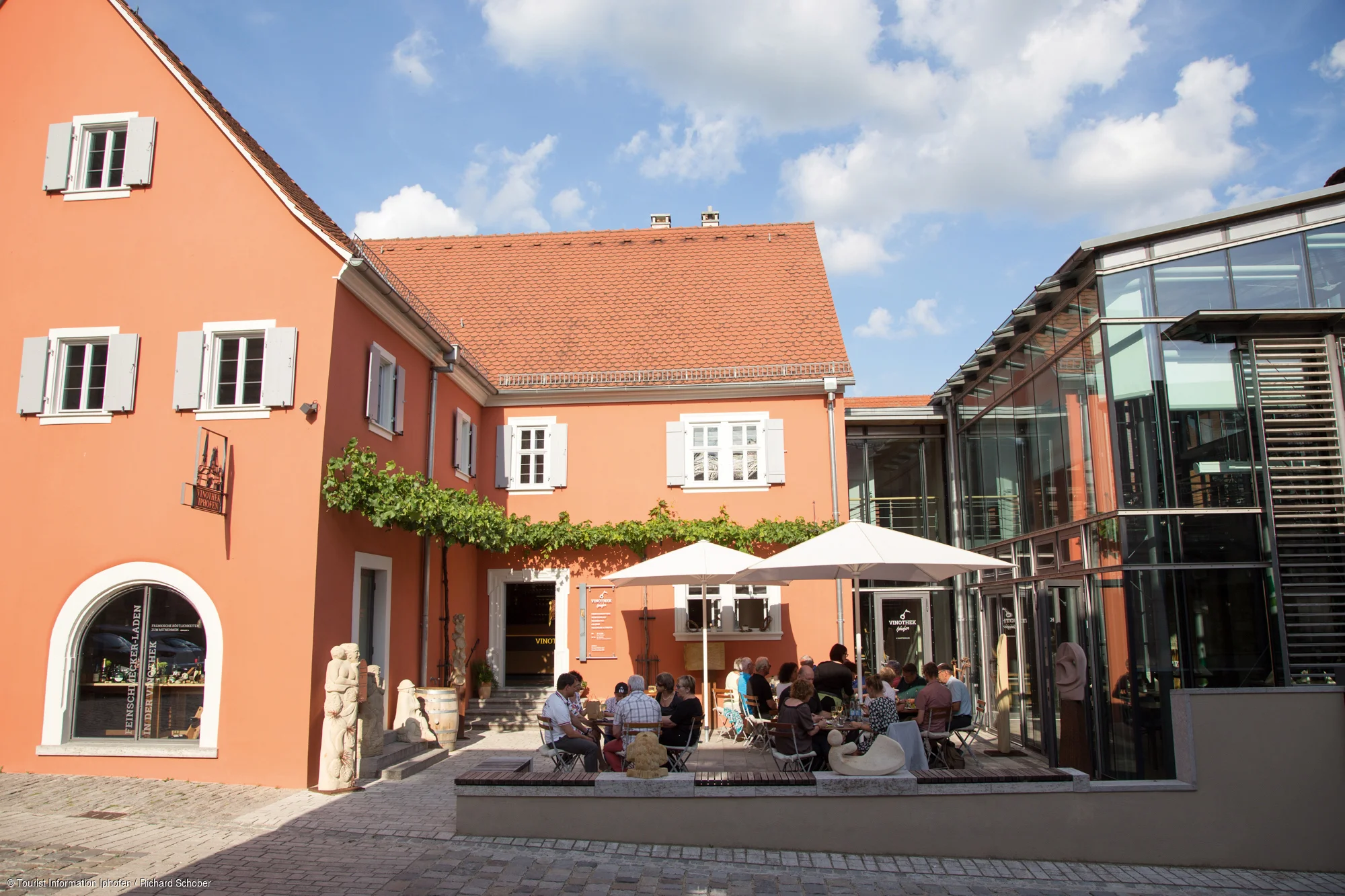 Innenhof mit orangem Gebäude, Glasfassade, Sitzbereich mit Tischen, Stühlen und Sonnenschirmen, Menschen sitzen draußen