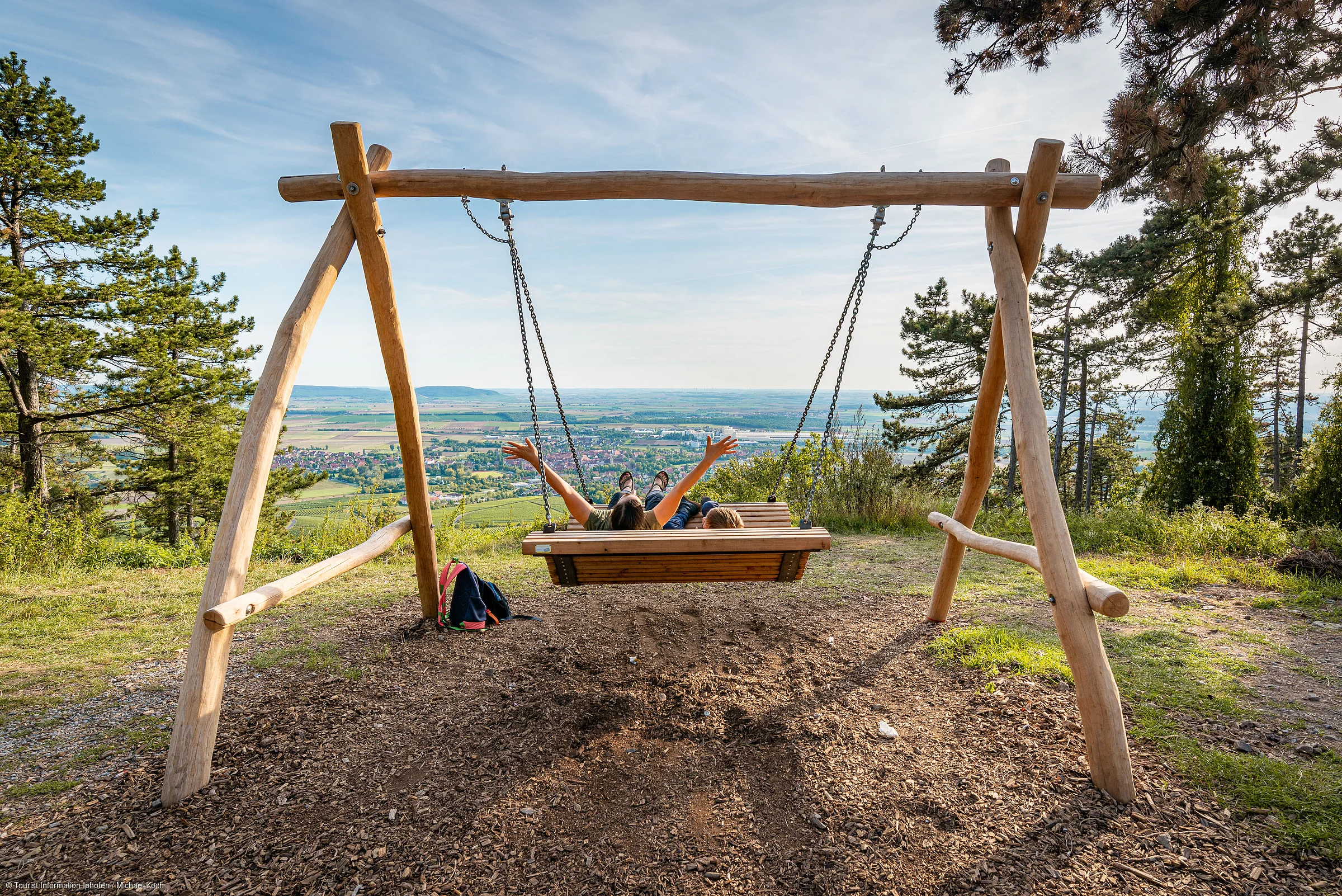 Zwei Personen sitzen auf einer großen Holzschaukel mit Blick auf ein Tal und eine Stadt im Hintergrund.