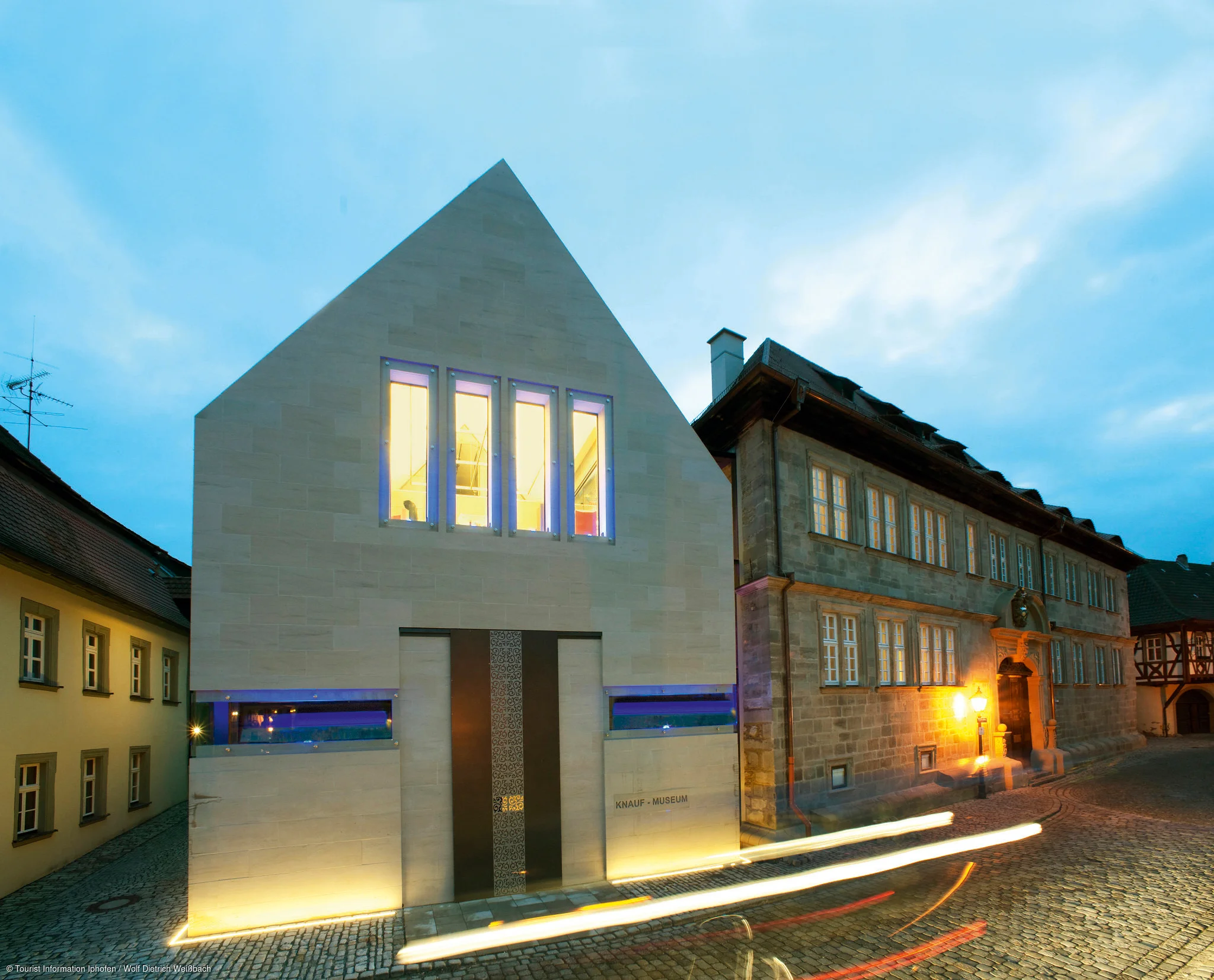 Abendaufnahme des modernen Knauf-Museums neben einem historischen Gebäude in einer gepflasterten Straße.