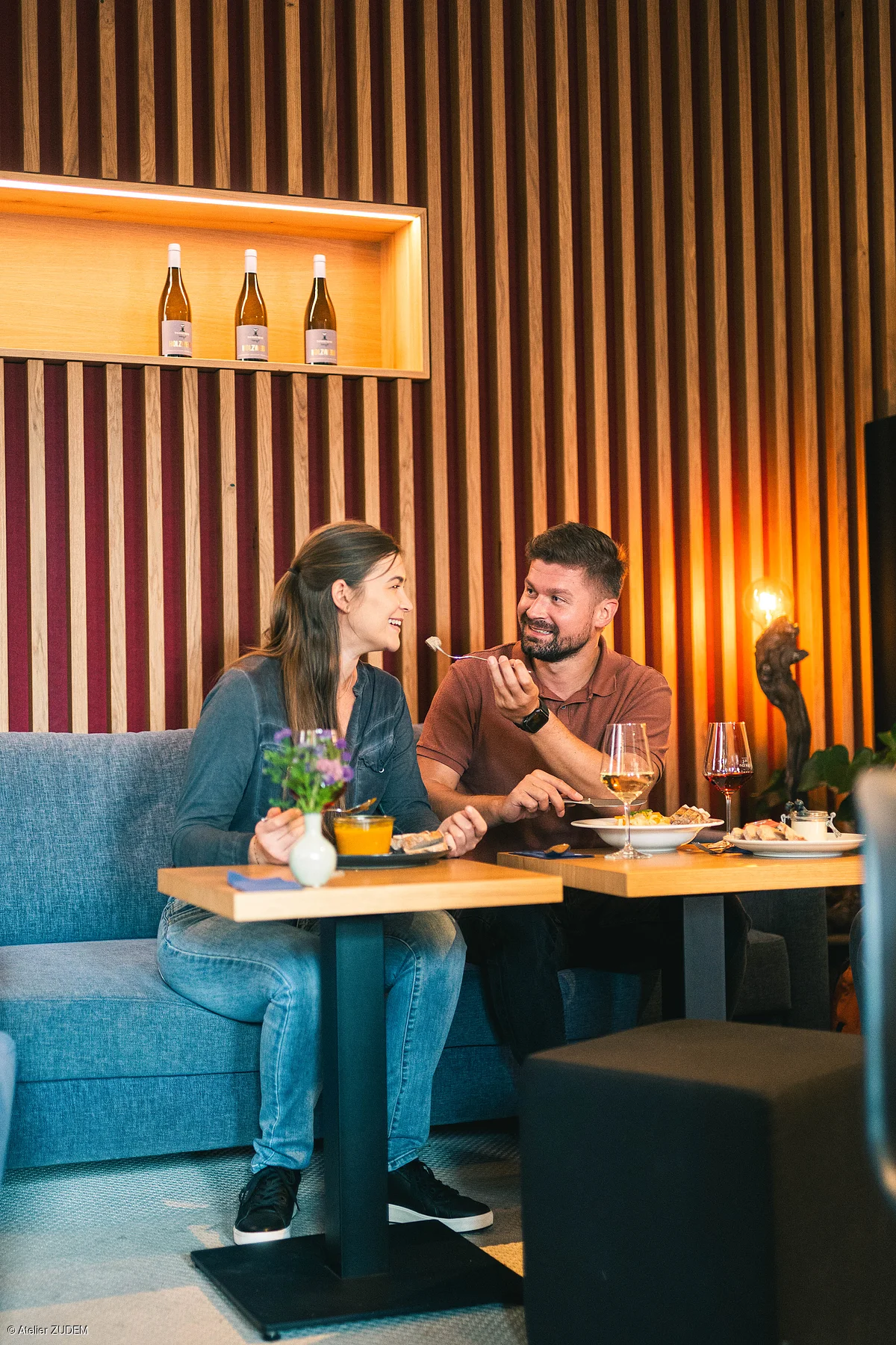 Zwei Personen sitzen an Tischen in einem Restaurant mit Holzpaneelwand und essen gemeinsam.