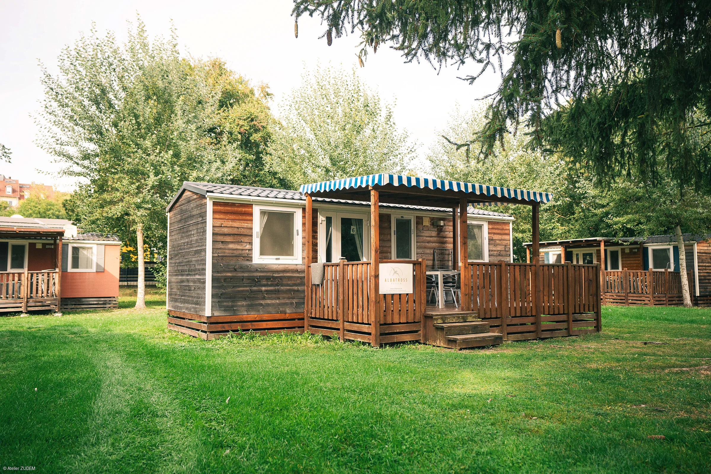 Holzhaus mit Veranda und blau-weiß gestreiftem Sonnenschutz auf grünem Rasen, umgeben von Bäumen.
