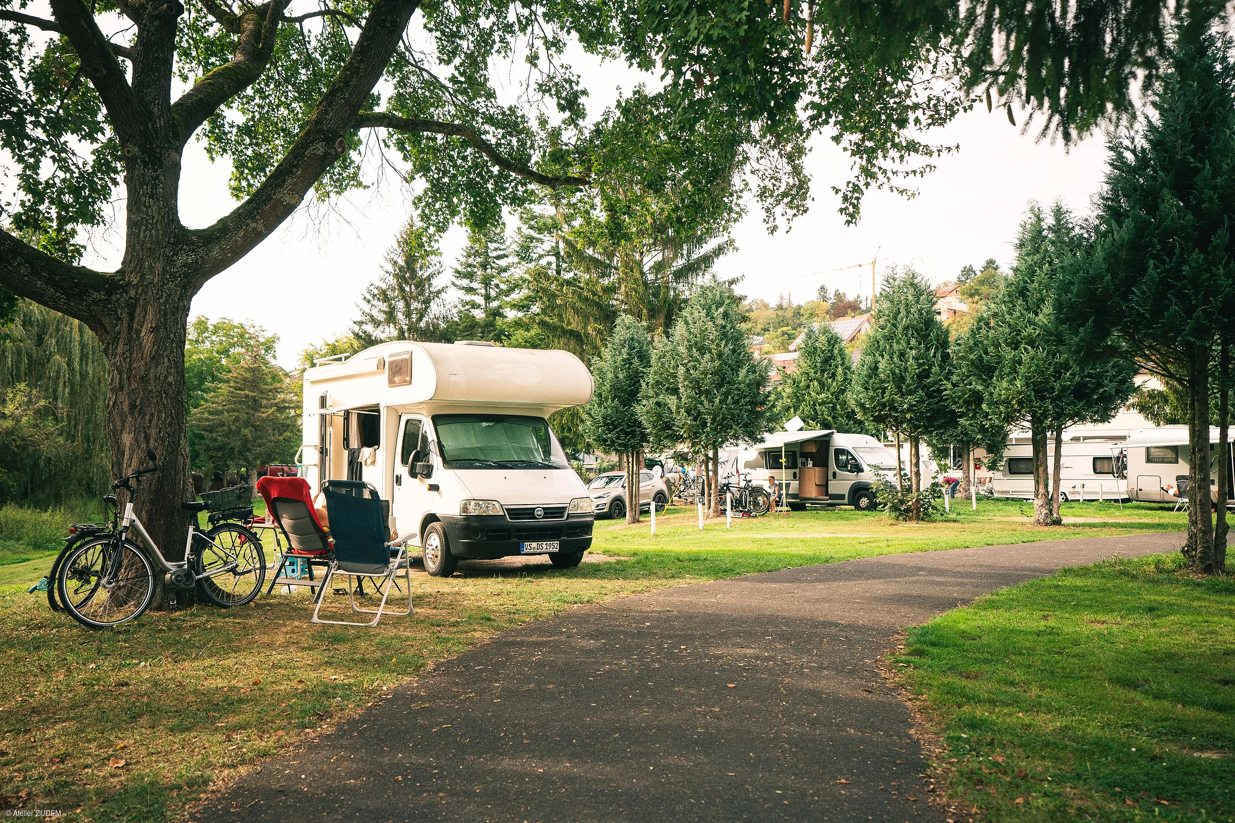 Campingplatz mit Wohnmobilen, Fahrrädern und Campingstühlen unter Bäumen neben einem Weg