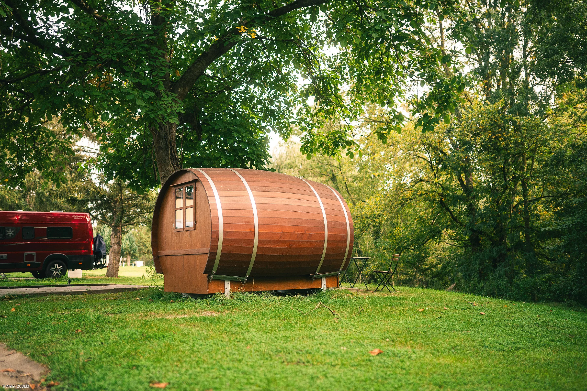 Holzhaus in Fassform auf einer Wiese unter Bäumen mit rotem Van im Hintergrund und Gartenstuhl mit Tisch