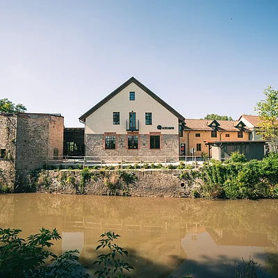 Gebäude mit Giebeldach und Steinmauer am Ufer eines Flusses unter blauem Himmel.
