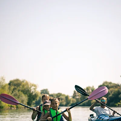 Drei Kinder in grünem Kajak und Mann in blauem Kajak paddeln auf ruhigem Fluss mit Bäumen im Hintergrund.