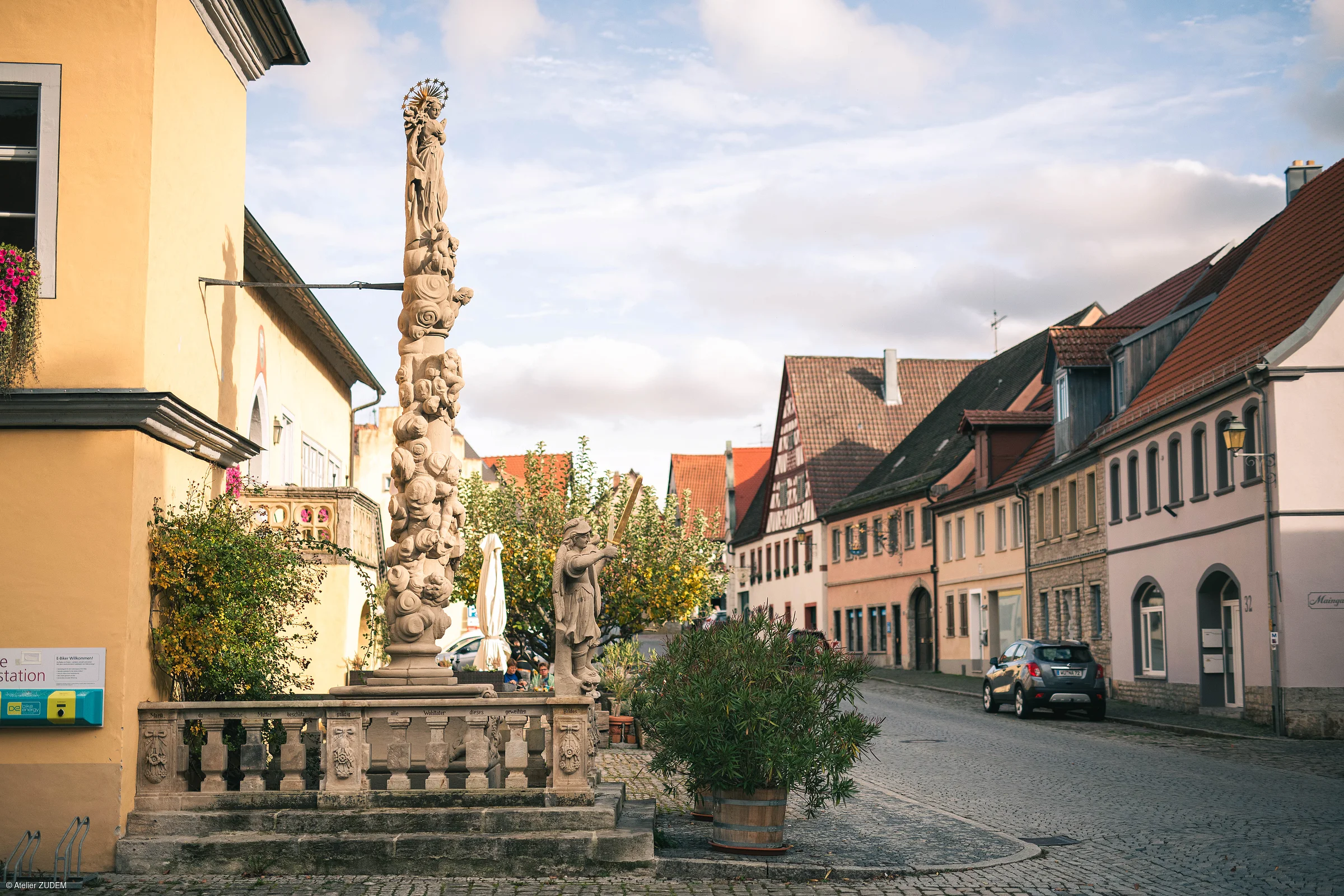 Steinerne Mariensäule und Statue an einer Straße mit Fachwerkhäusern und parkenden Autos bei Tageslicht.