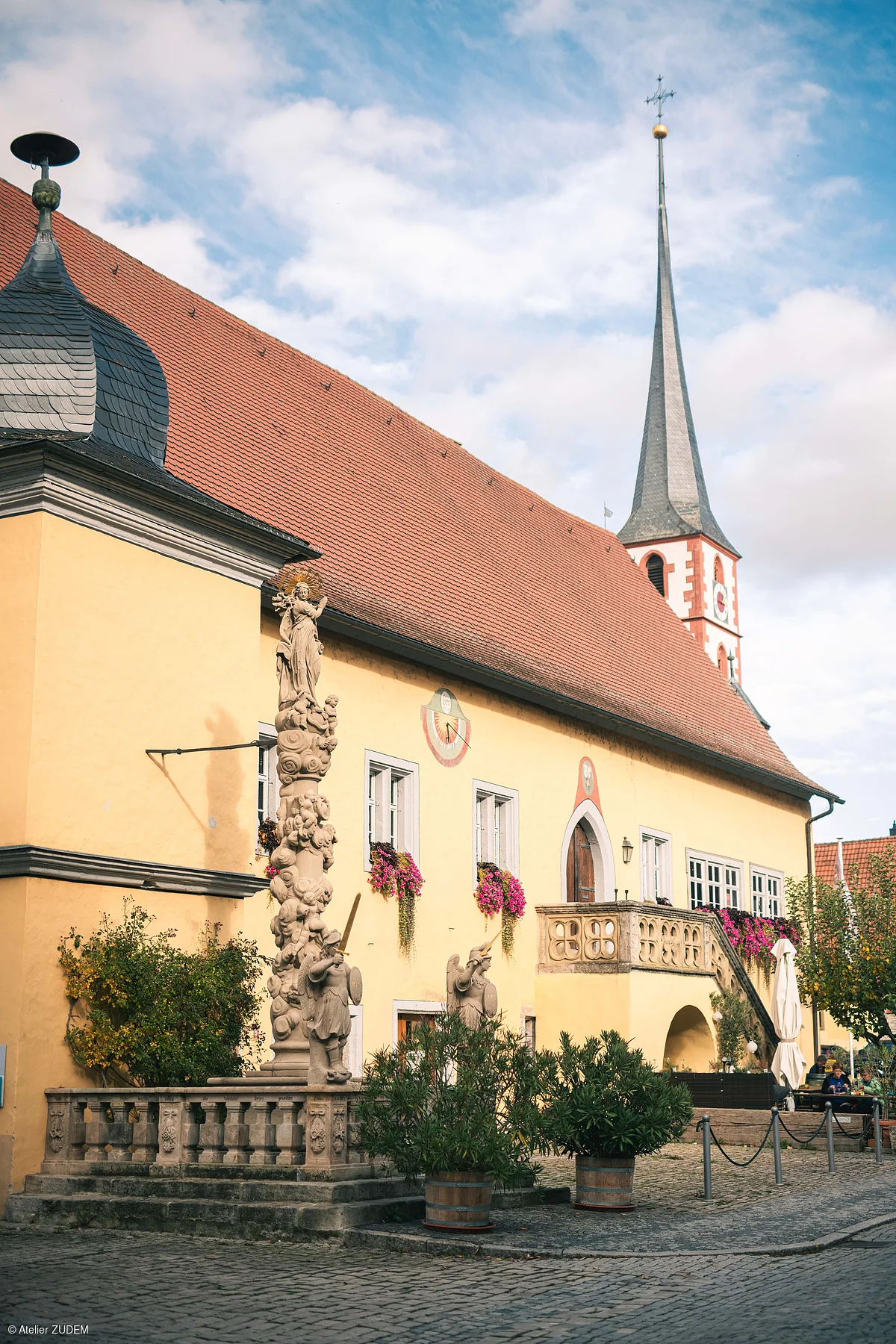 Kirchengebäude mit rotem Dach, Turm mit Kreuz, Sonnenuhr und Mariensäule vor gelber Fassade bei Tageslicht