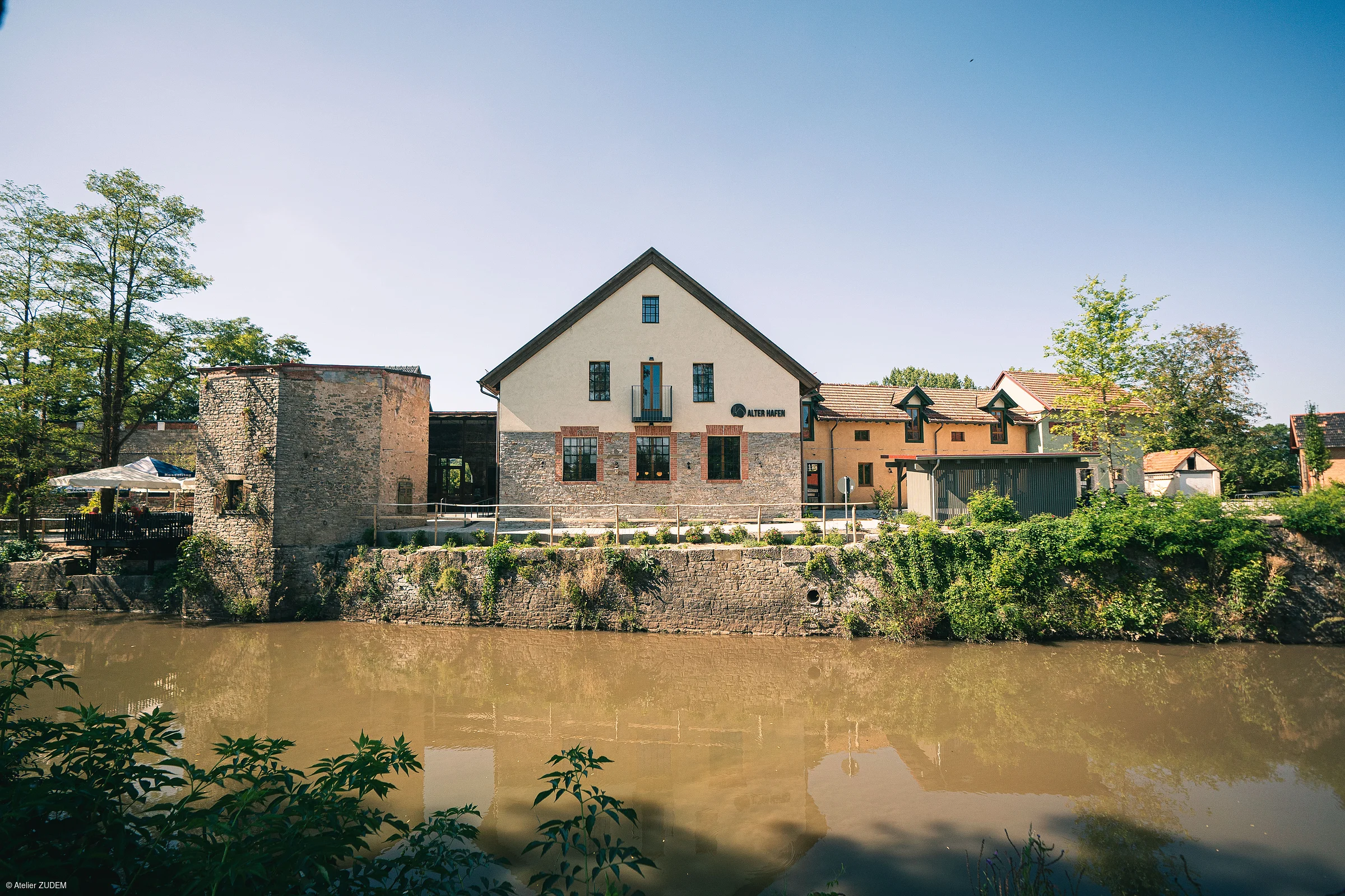 Gebäude mit Giebeldach und Steinmauer am Ufer eines Flusses unter blauem Himmel.