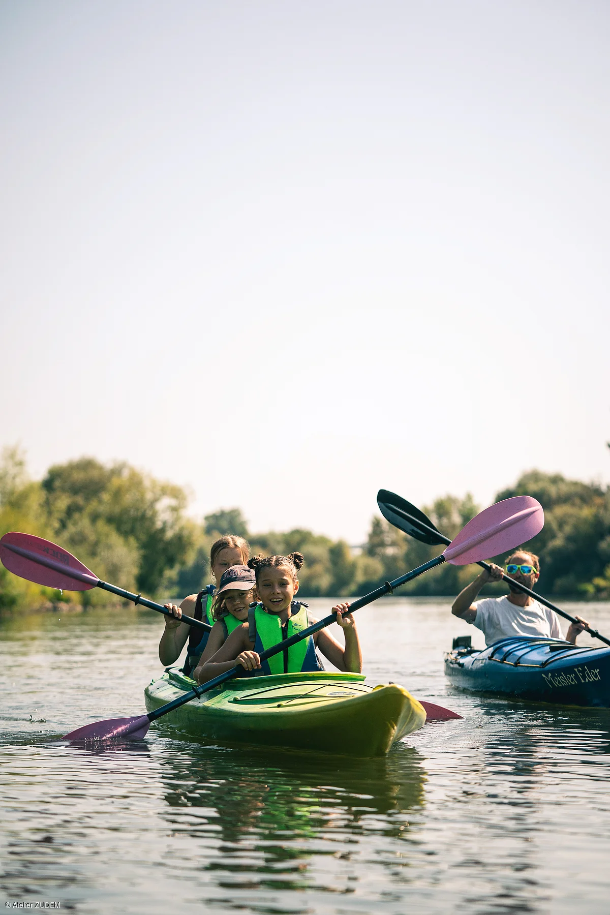 Drei Kinder in grünem Kajak und Mann in blauem Kajak paddeln auf ruhigem Fluss mit Bäumen im Hintergrund.