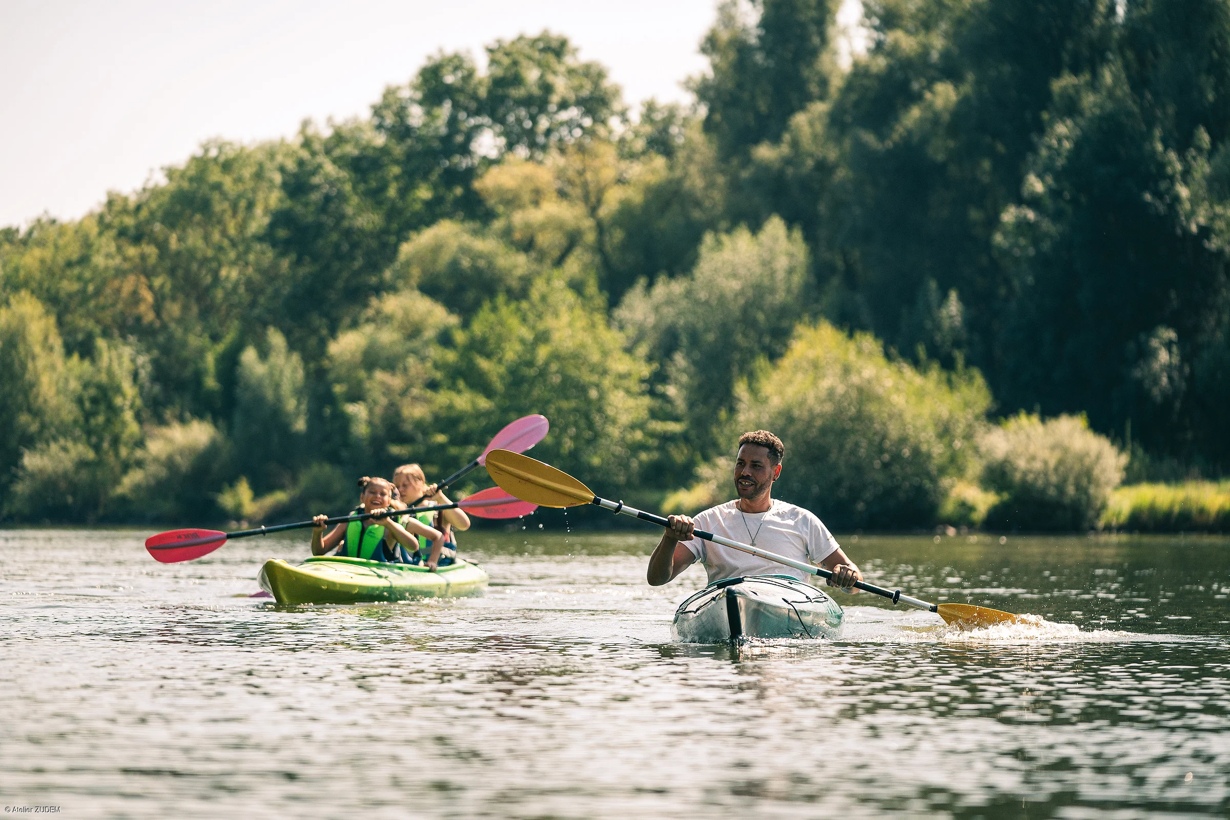 Mann und zwei Kinder paddeln mit Kajaks auf einem Fluss vor bewaldetem Ufer bei Tageslicht.