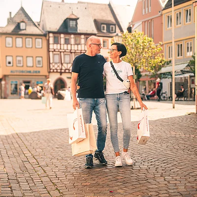 Älteres Paar mit Einkaufstüten geht Arm in Arm über gepflasterte Straße in Altstadt bei Sonnenschein.