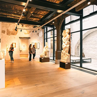 Innenraum eines Museums mit Holzdecke, großen Fenstern, Skulpturen und drei Personen, die Kunstwerke betrachten.