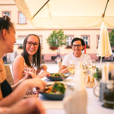 Vier Personen sitzen lachend an einem gedeckten Tisch im Freien unter einem Sonnenschirm und essen.