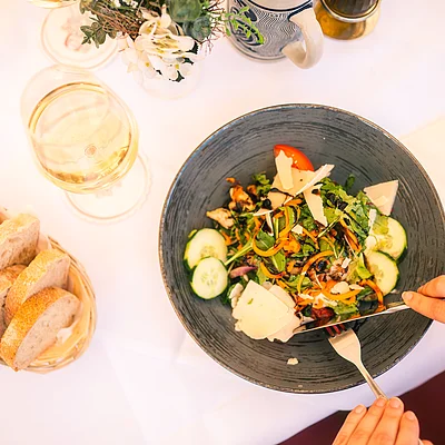 Person schneidet Salat mit Käse in grauer Schüssel, daneben Brotkorb und Glas Weißwein auf weißer Tischdecke