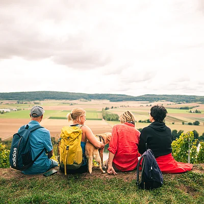 Vier Personen mit Rucksäcken und ein Hund sitzen auf einer Wiese mit Blick auf Felder und Hügel.