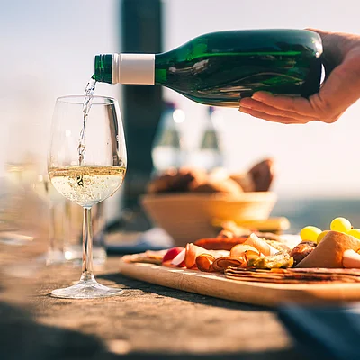 Weißwein wird aus einer grünen Flasche in ein Weinglas gegossen, daneben liegt eine Platte mit Aufschnitt und Trauben.