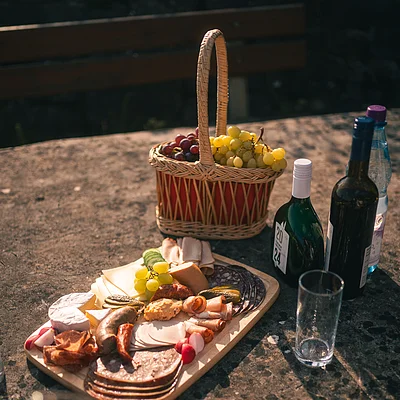 Brett mit verschiedenen Wurst- und Käsesorten, Traubenkorb, Weinflaschen und Glas auf Steintisch im Freien.