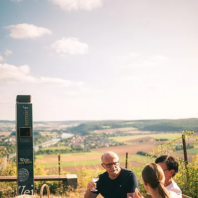 Drei Personen sitzen an einem Tisch mit Wein und Snacks, im Hintergrund Landschaft mit Feldern und Dorf.