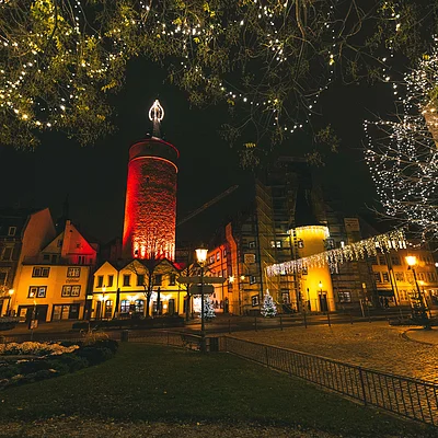 Nachtaufnahme eines beleuchteten Stadtplatzes mit altem Turm, Häusern und Weihnachtsbeleuchtung in Bäumen.