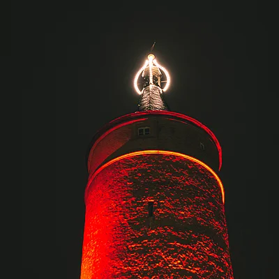 Turm mit roter Beleuchtung und leuchtendem Ring an der Spitze bei Nacht vor dunklem Himmel.