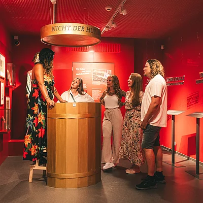 Fünf Personen in einem roten Ausstellungsraum mit Holzfass.
