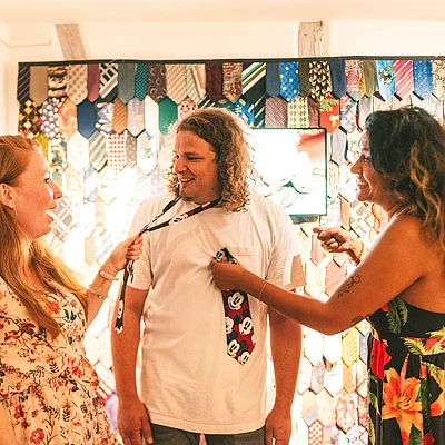 Drei Personen lachen, zwei Frauen halten Krawatten, im Hintergrund Wand mit vielen bunten Krawatten.