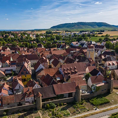 Luftaufnahme einer Stadt mit roten Dächern, einer Stadtmauer und einem Turm vor Hügeln und Feldern im Hintergrund.