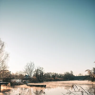 Fluss mit Nebel, kahlen Bäumen, Boot und Brücke bei klarem Himmel im Winter.
