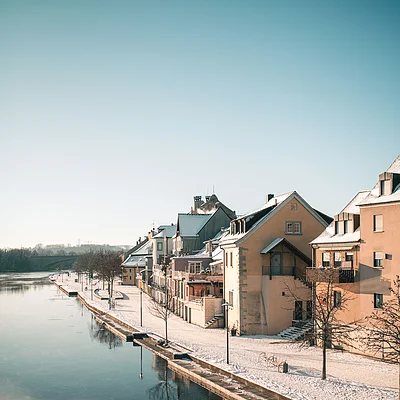 Schneebedeckte Häuser und Uferpromenade an einem ruhigen Fluss unter klarem Himmel im Winter.