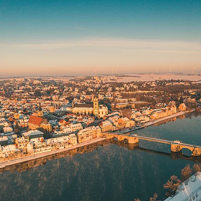 Luftaufnahme einer verschneiten Stadt mit Fluss und Brücke bei Sonnenaufgang oder Sonnenuntergang.