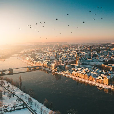 Vogelperspektive auf eine verschneite Stadt mit Fluss, Brücke und Vögeln im Sonnenaufgang.