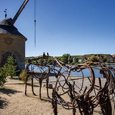Skulptur aus Metallstäben in Pferdeform vor rundem Steingebäude und Flussufer mit Häusern im Hintergrund bei klarem Himmel.