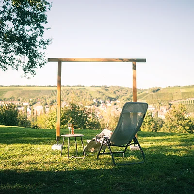 Person sitzt auf Liegestuhl mit Tisch und Getränk, im Hintergrund Weinberge und Dorf sichtbar
