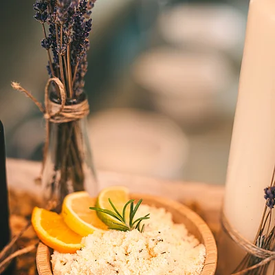 Holzschale mit Badesalz, Zitronen- und Orangenscheiben, daneben getrockneter Lavendel in Glasflasche und Kerze.