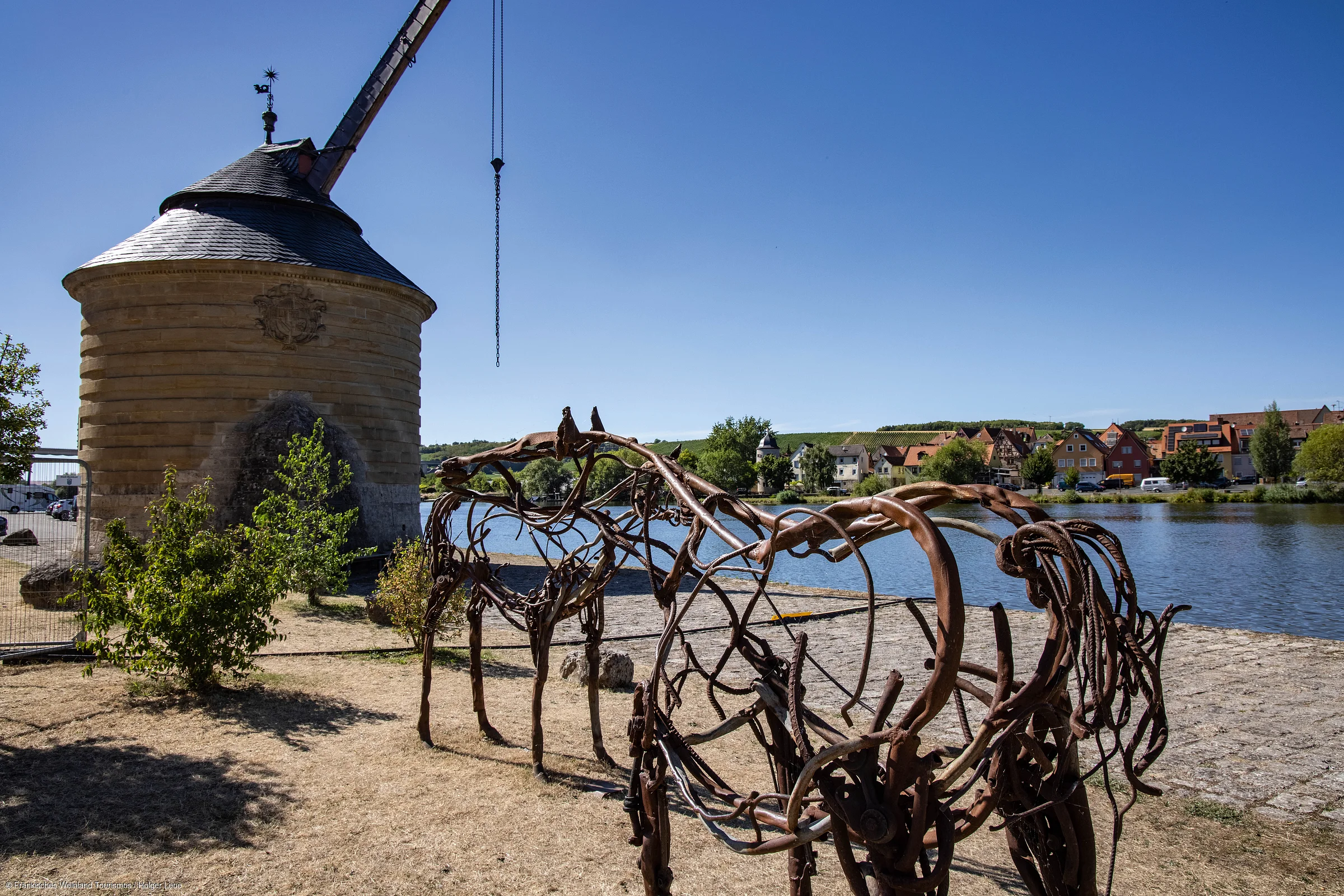 Skulptur aus Metallstäben in Pferdeform vor rundem Steingebäude und Flussufer mit Häusern im Hintergrund bei klarem Himmel.