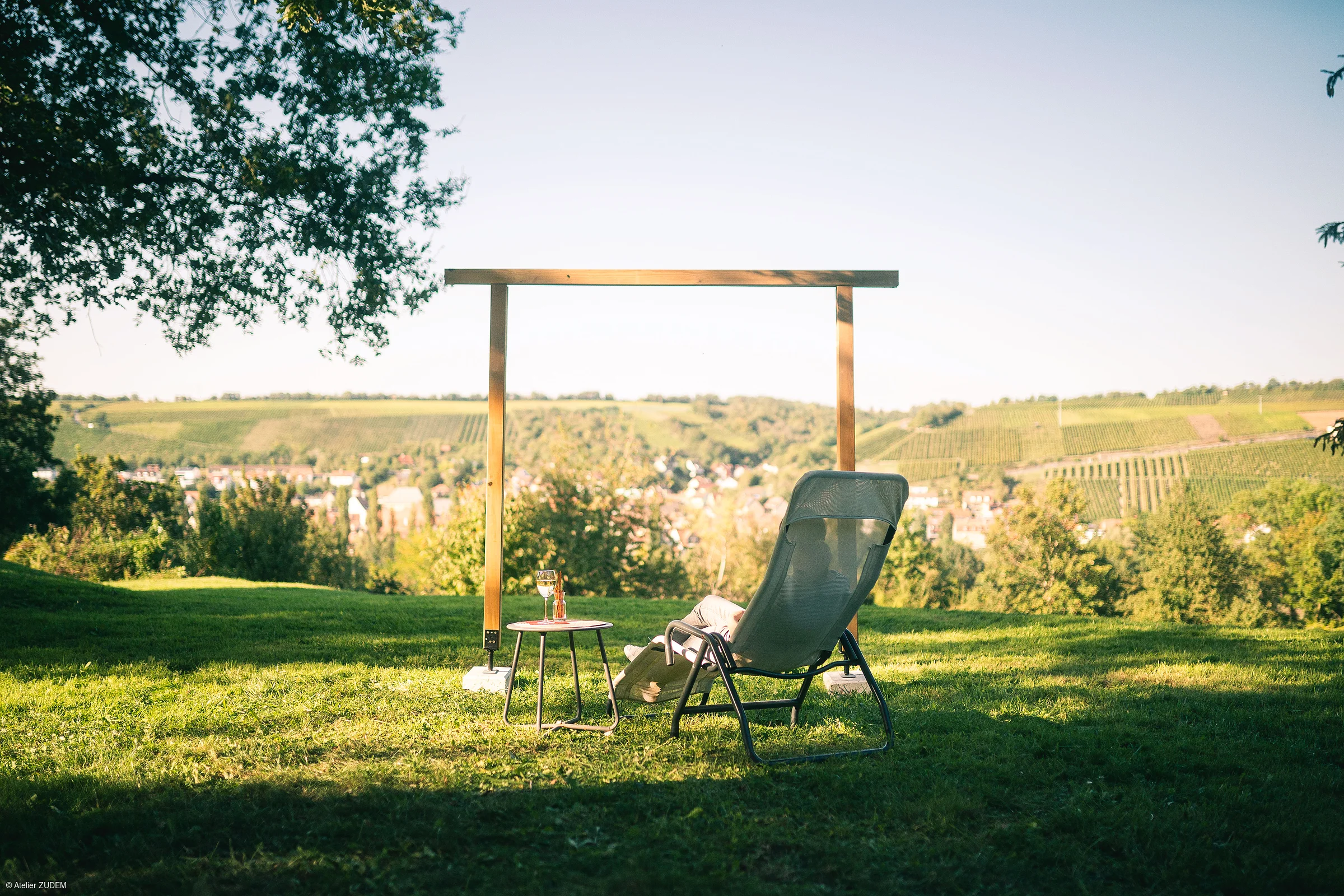 Person sitzt auf Liegestuhl mit Tisch und Getränk, im Hintergrund Weinberge und Dorf sichtbar