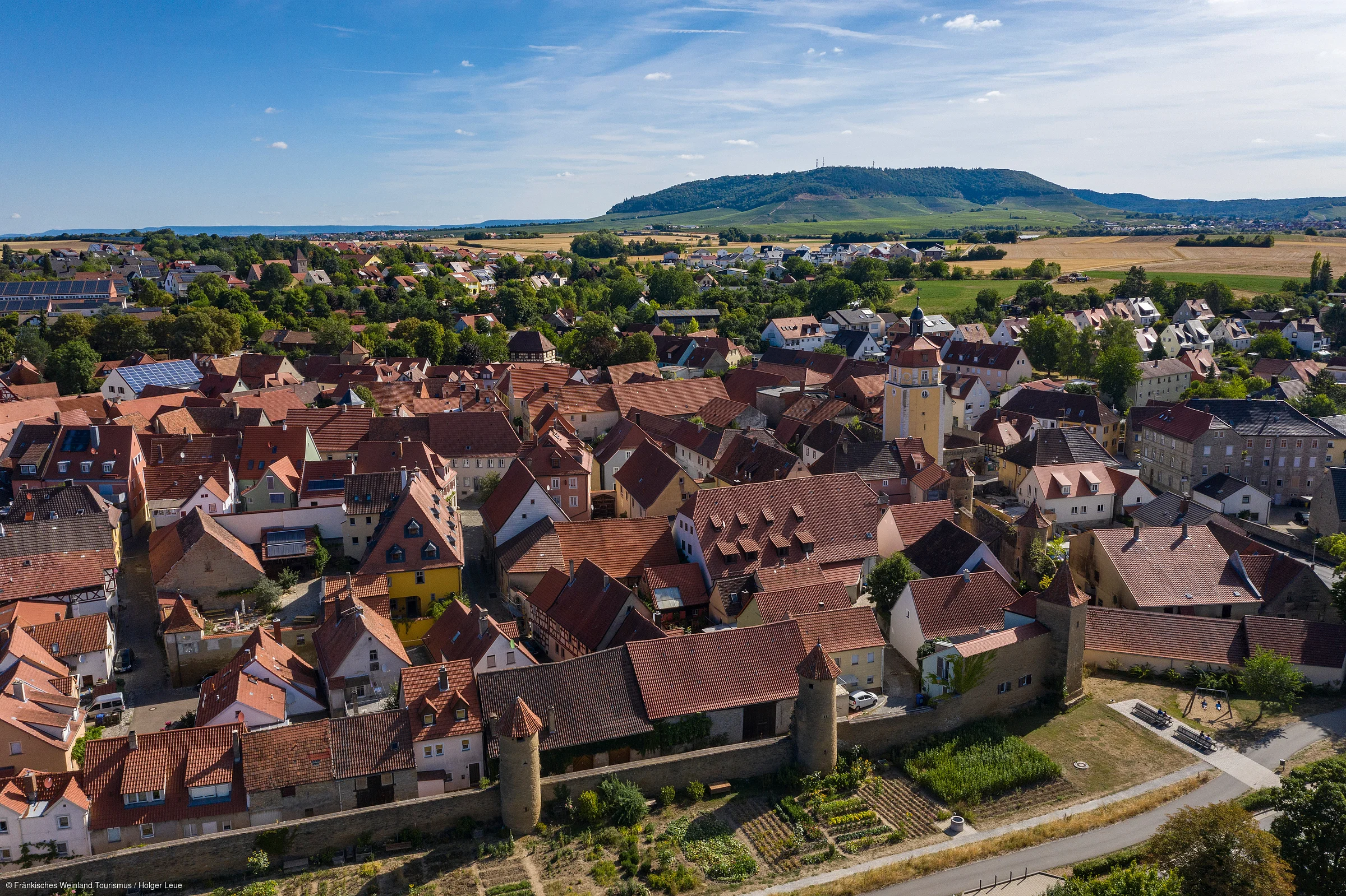 Luftaufnahme einer Stadt mit roten Dächern, einer Stadtmauer und einem Turm vor Hügeln und Feldern im Hintergrund.