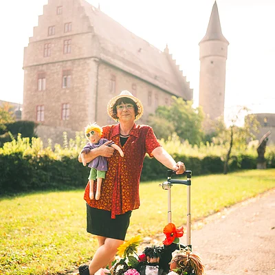 Frau mit Strohhut hält Puppe und steht neben Rollator mit Blumenkorb vor historischem Gebäude.