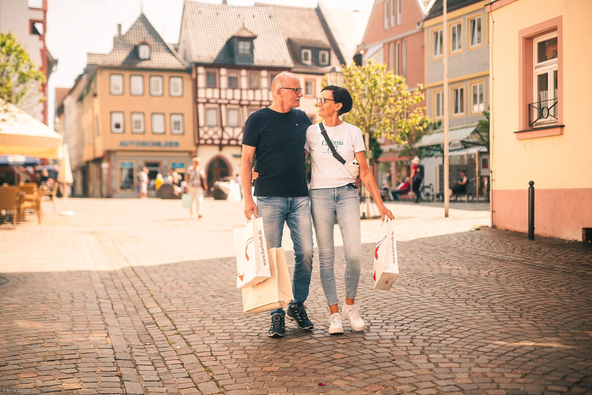 Älteres Paar mit Einkaufstüten geht Arm in Arm über gepflasterte Straße in Altstadt bei Sonnenschein.