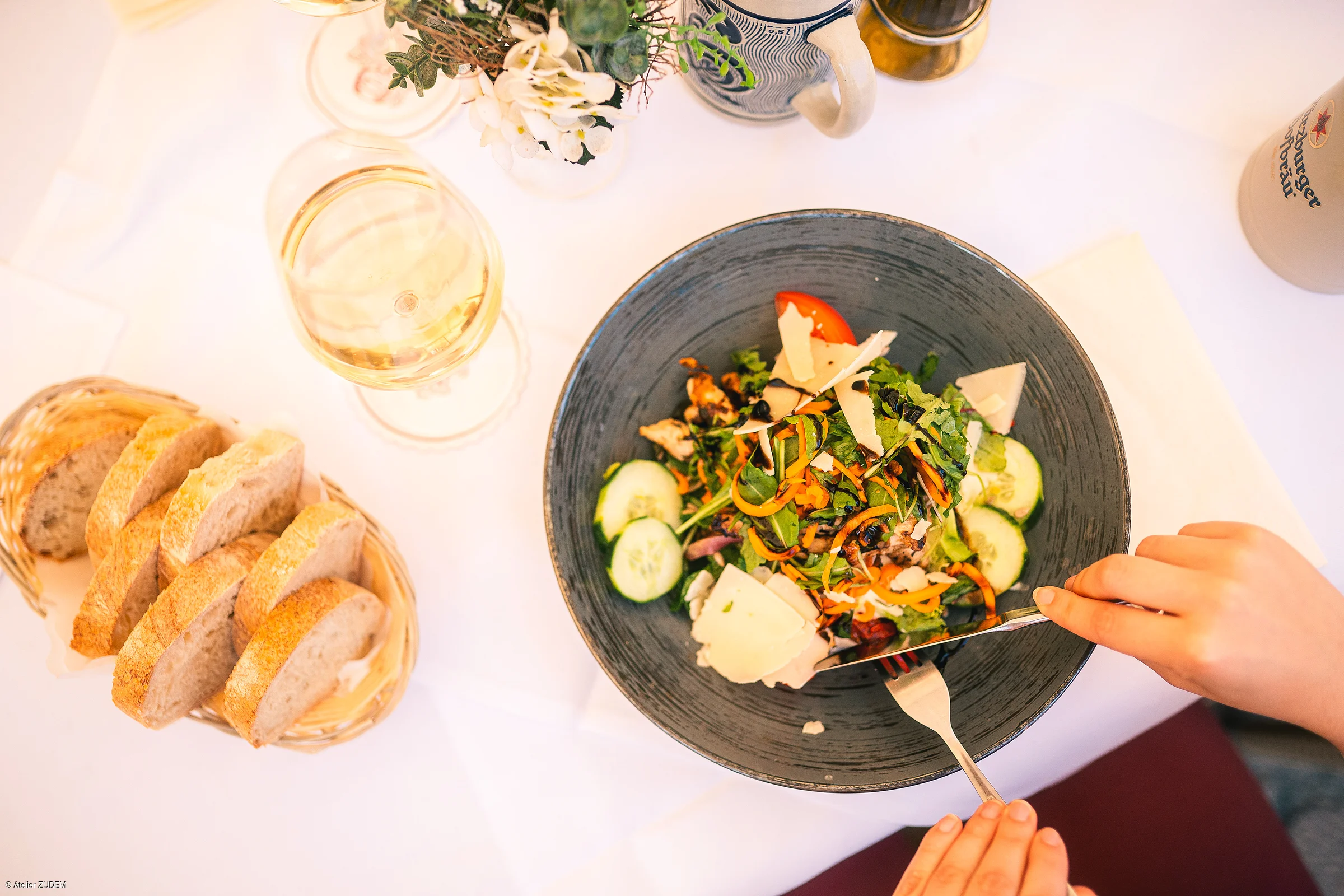 Person schneidet Salat mit Käse in grauer Schüssel, daneben Brotkorb und Glas Weißwein auf weißer Tischdecke