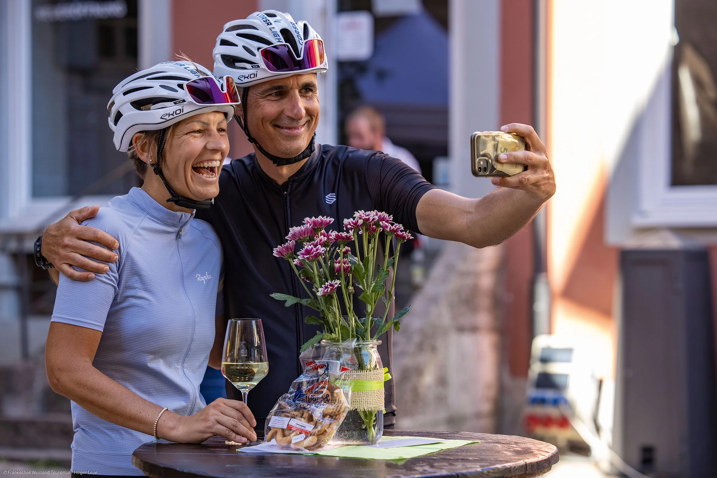 Zwei Radfahrer mit Helmen machen lachend ein Selfie an einem Tisch mit Blumen und Snacks.