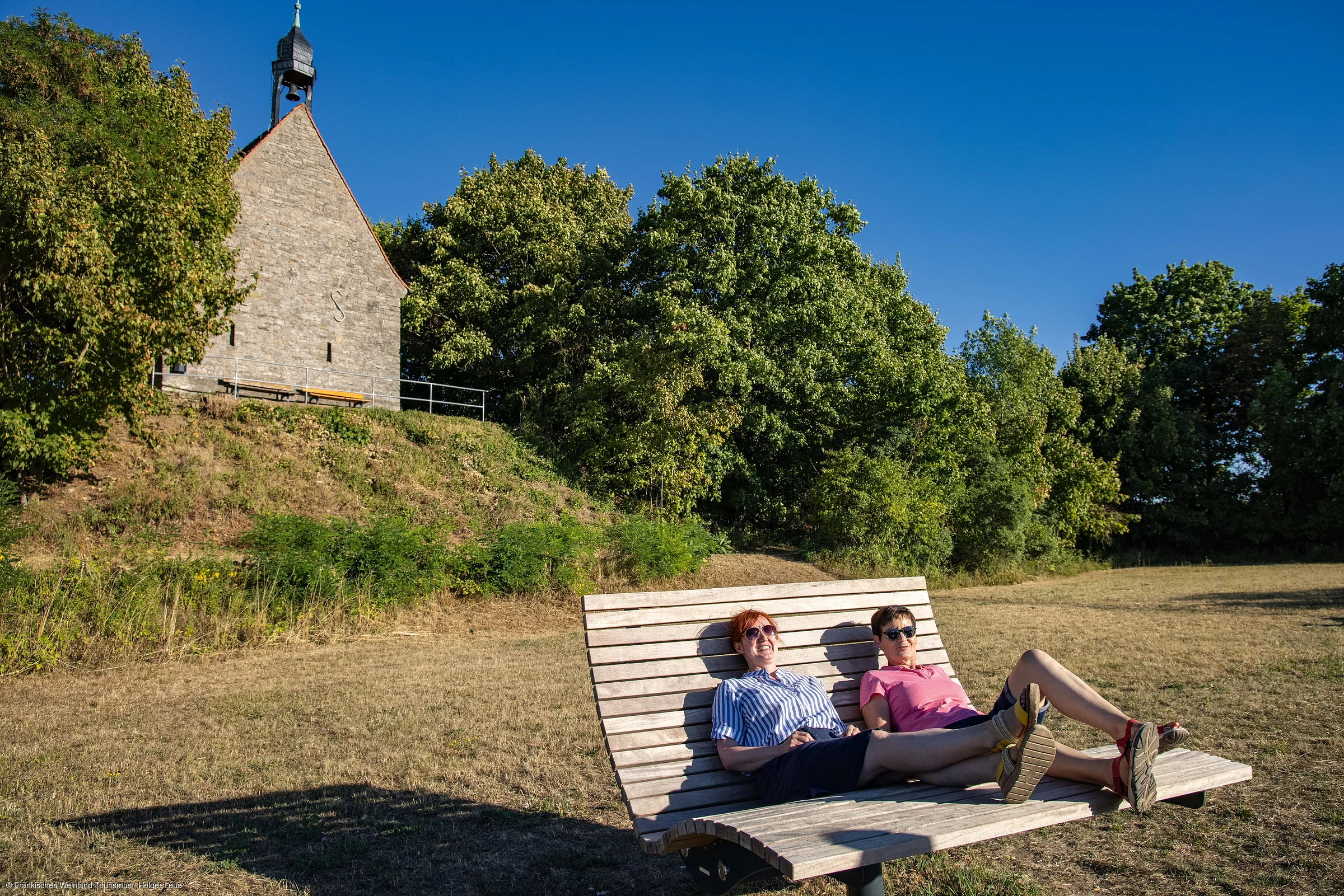 Zwei Frauen liegen auf einer großen Holzliege auf einer Wiese, im Hintergrund Bäume und ein Steinhaus mit Glockenturm.